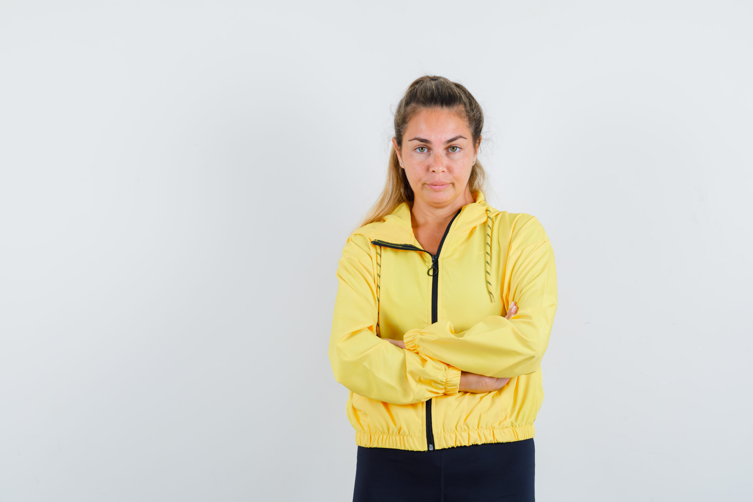 blonde woman in yellow bomber jacket and black pants standing arms crossed and looking happy , front view.